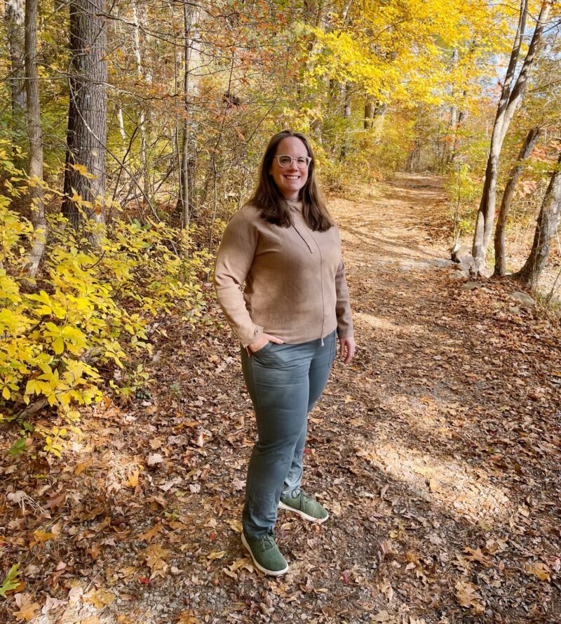 Eileen Cotter Wright of Pure Wander outside in the New England fall forest with yellow leaves surrounding her in sneakers, grey pants and a quince hoodie eileen cotter wright